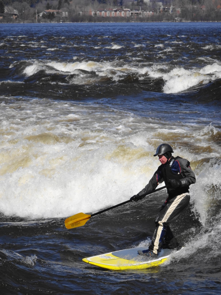 A stand-up paddleboarder takes his place on the "The Wave".  More whitewater lies in the midground of the photograph, while the Quebec shoreline lies in the background.