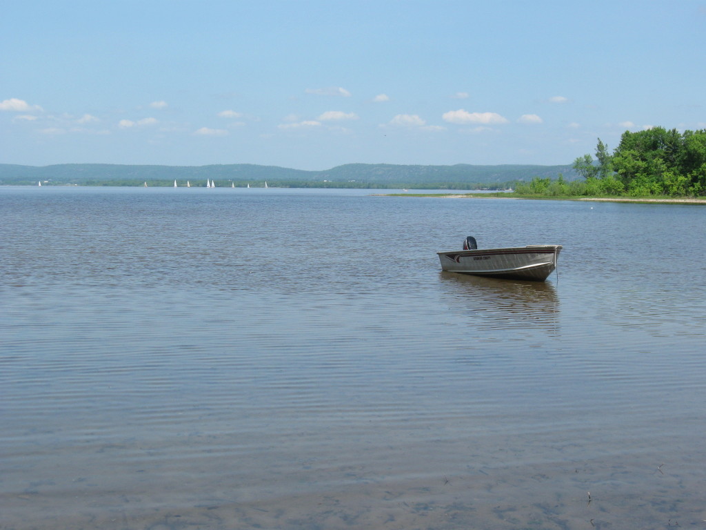 An aluminum boat floats in the shallows of Constance Bay at the end of Greenland Road, with sail boats and the Quebec shoreline in the distant background.