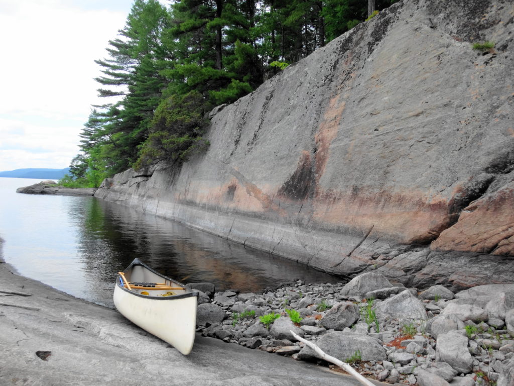 A canoe rests on a rocky shore in a small cove under a flat wall of stone.