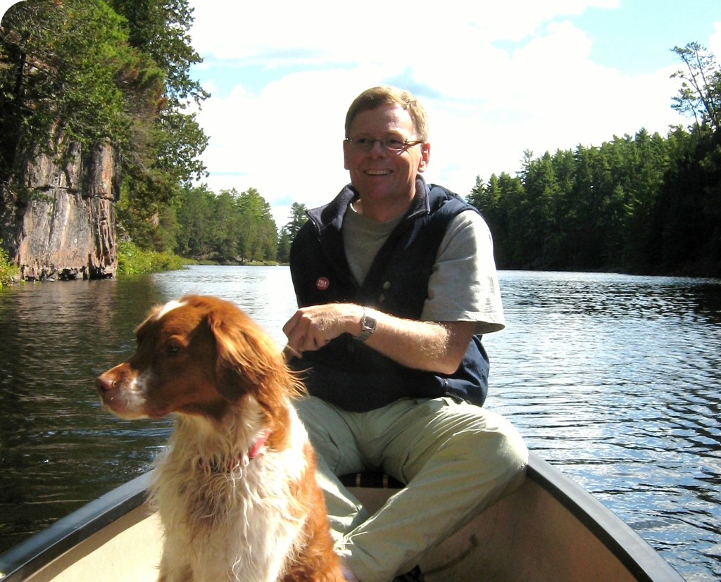 The author paddles his canoe along a river, with a beautiful Brittany Spaniel sitting up in the canoe.