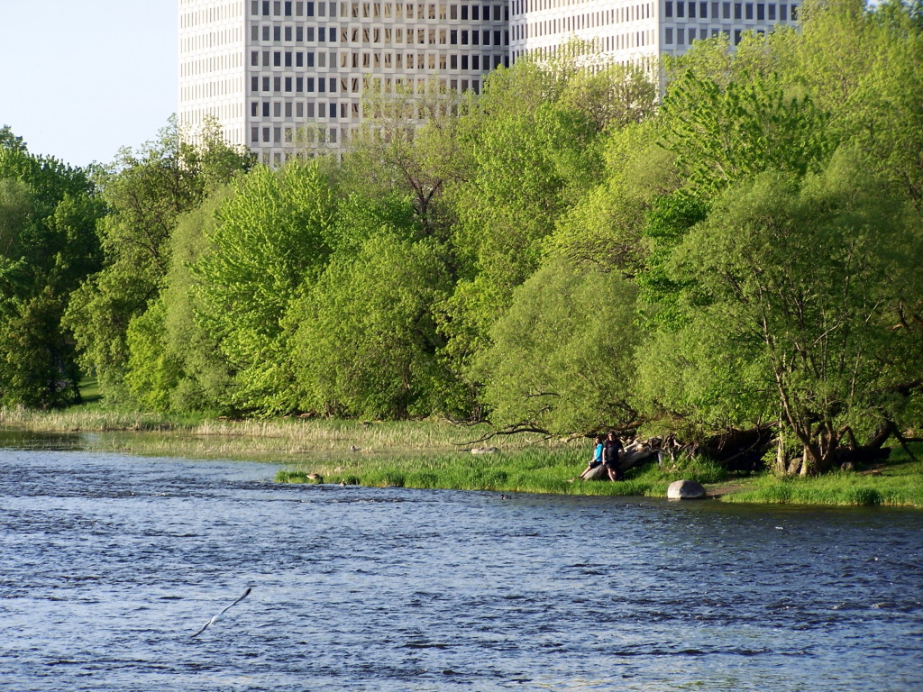 A young couple sit together on a log on the far, wooded shore of the river. Several towers loom in the distance.