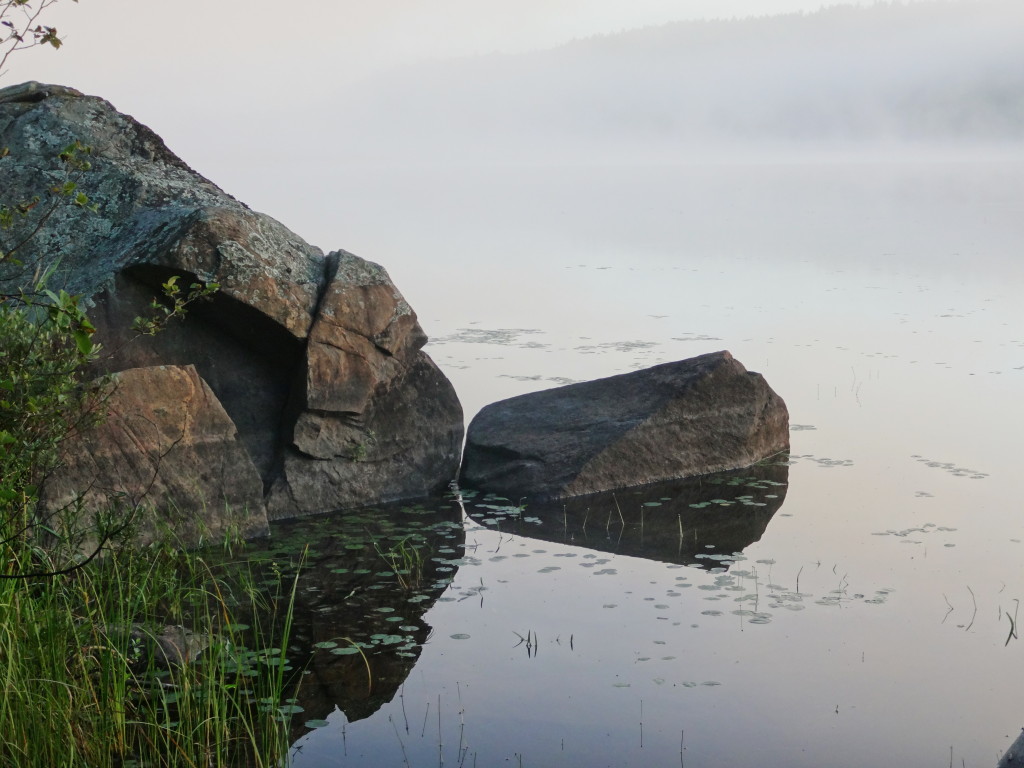 Boulders reflect in the still water of a mist covered lake in early morning light.
