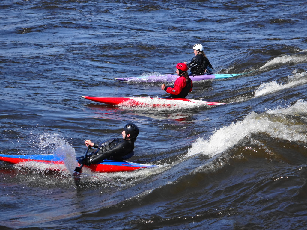 Three whitewater canoes slice down a standing wave.  Their canoes and their paddles throw up spray in the swiftly moving water.