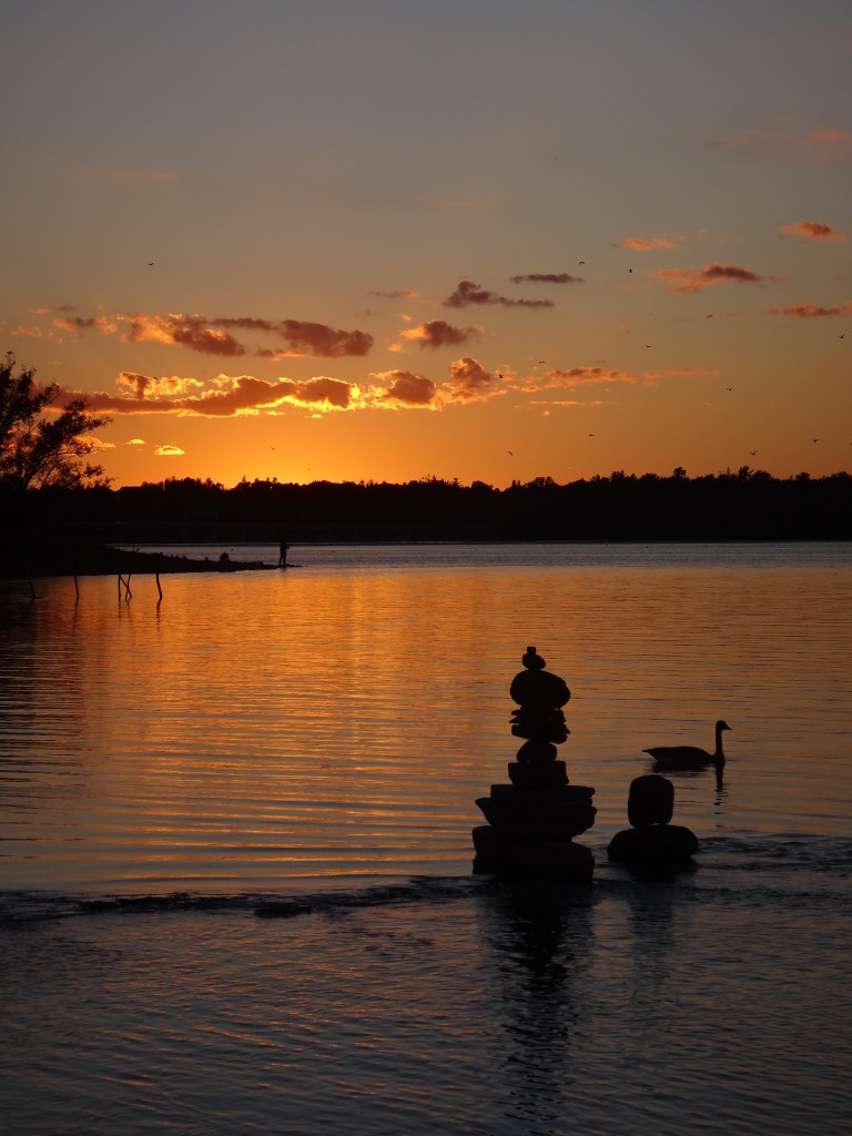 In the foreground, a balanced stone sculpture and a goose stand silhouetted against the glowing water of the river, while a fisherman stands in silhouette on a point in the distance.
