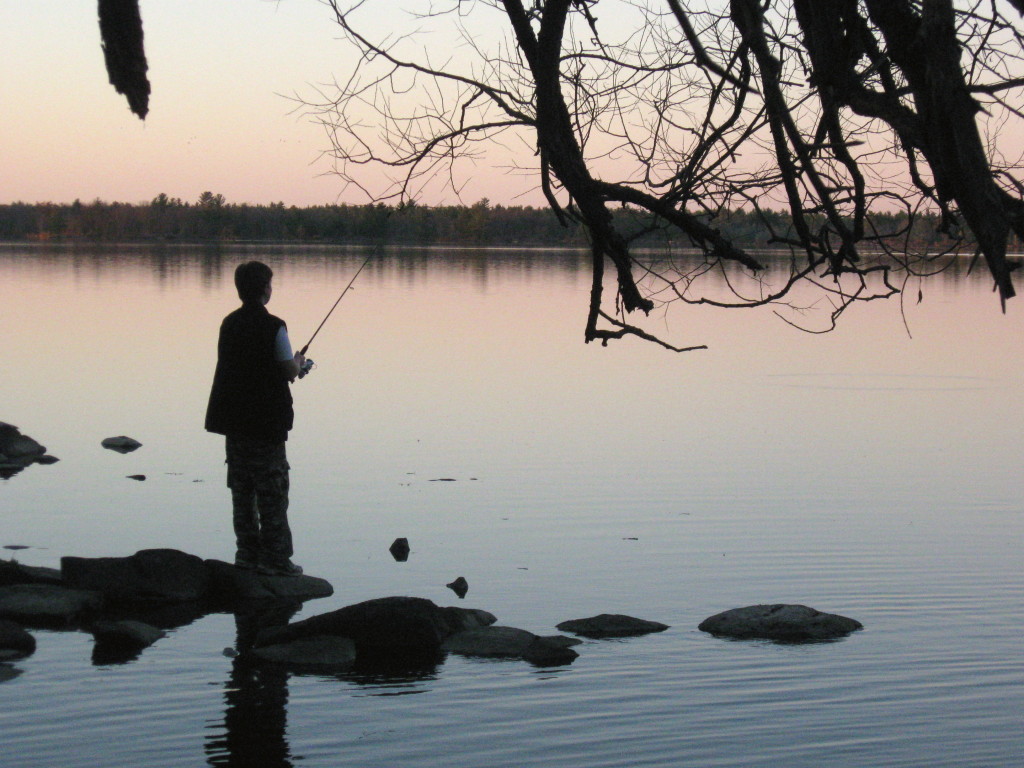 A teenage boy fishes from the shoreline, silhouetted against the river and sunset.