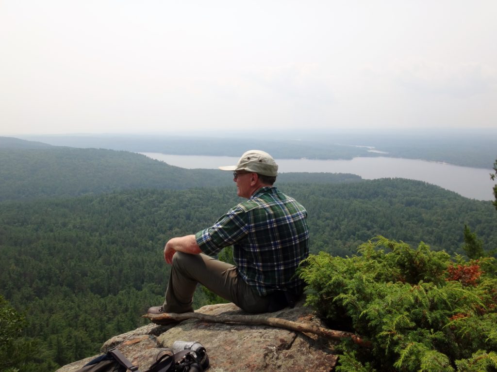 The author rests on a rocky lookout at the top of Mount Martin, with forest and the Ottawa River in the background.